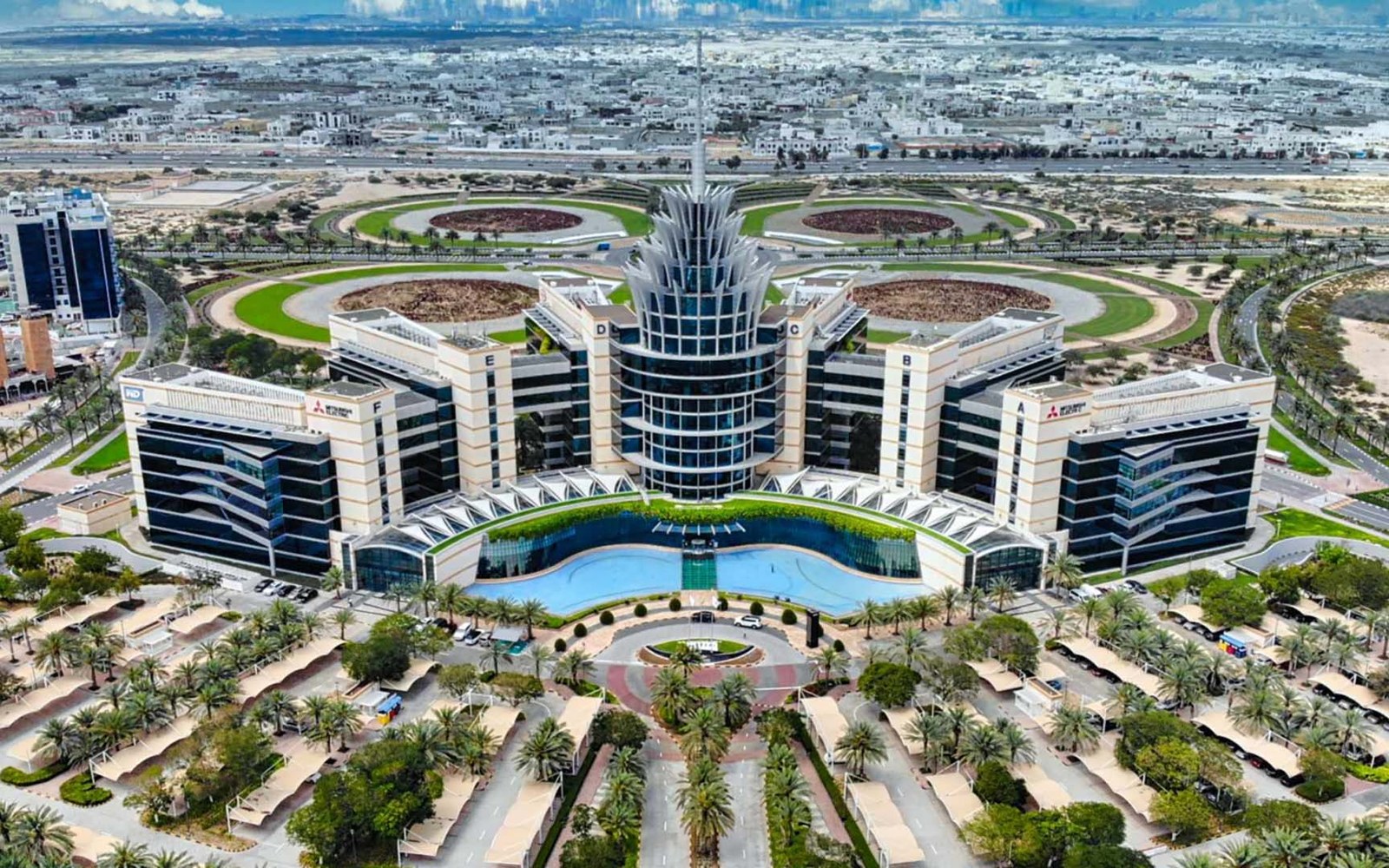 Aerial view of the Dubai Silicon Oasis headquarters building, with its spired central tower, blue pool and landscaped roundabouts, and residential neighbourhoods in the distance